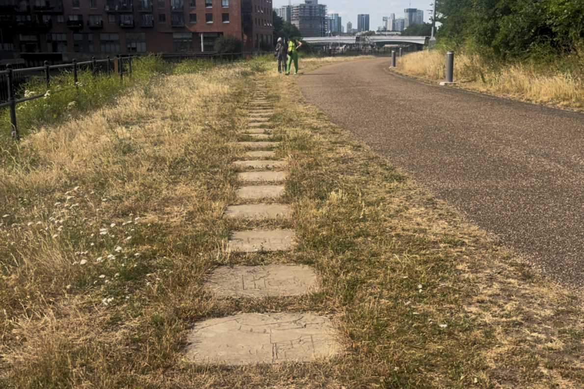 A grassy path with engraved stones runs alongside an paved trail, bordered by dry grass. Two people stand in the distance next to tall urban buildings.