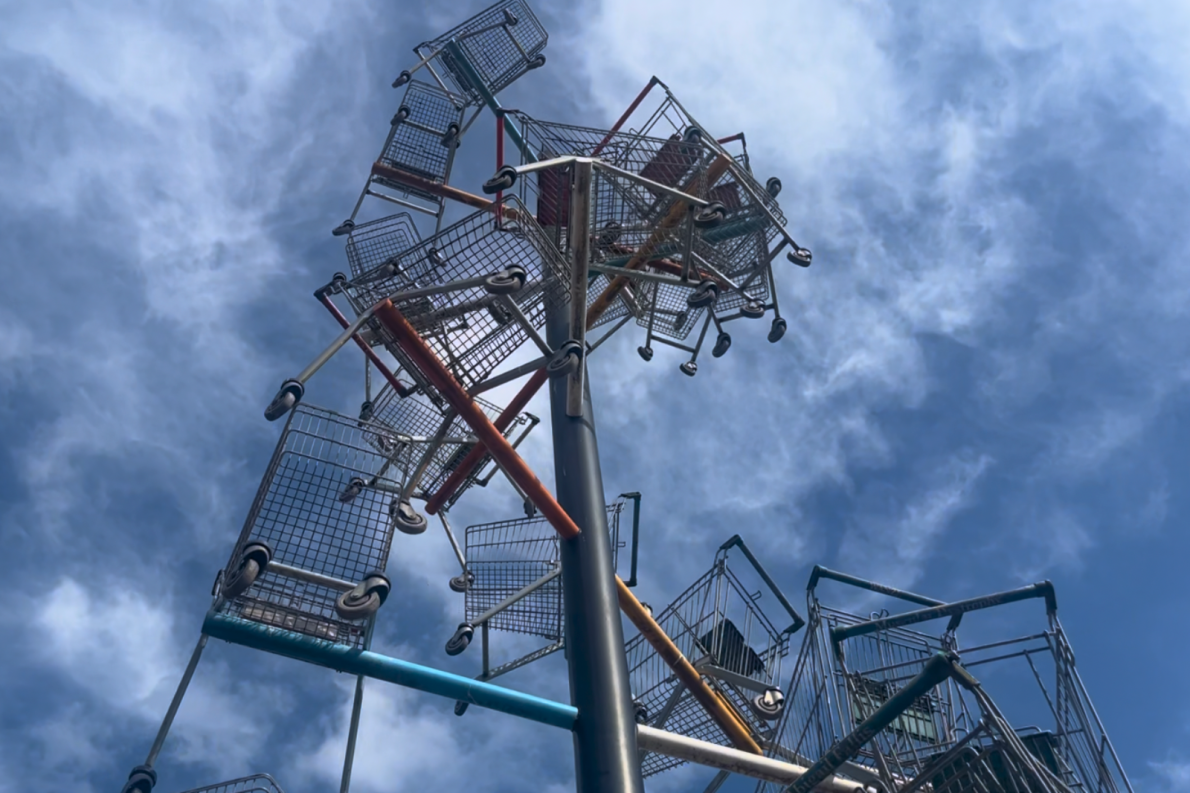 A sculpture of shopping carts stacked on metal poles, reaching toward a cloudy blue sky. The carts form an abstract, futuristic structure.
