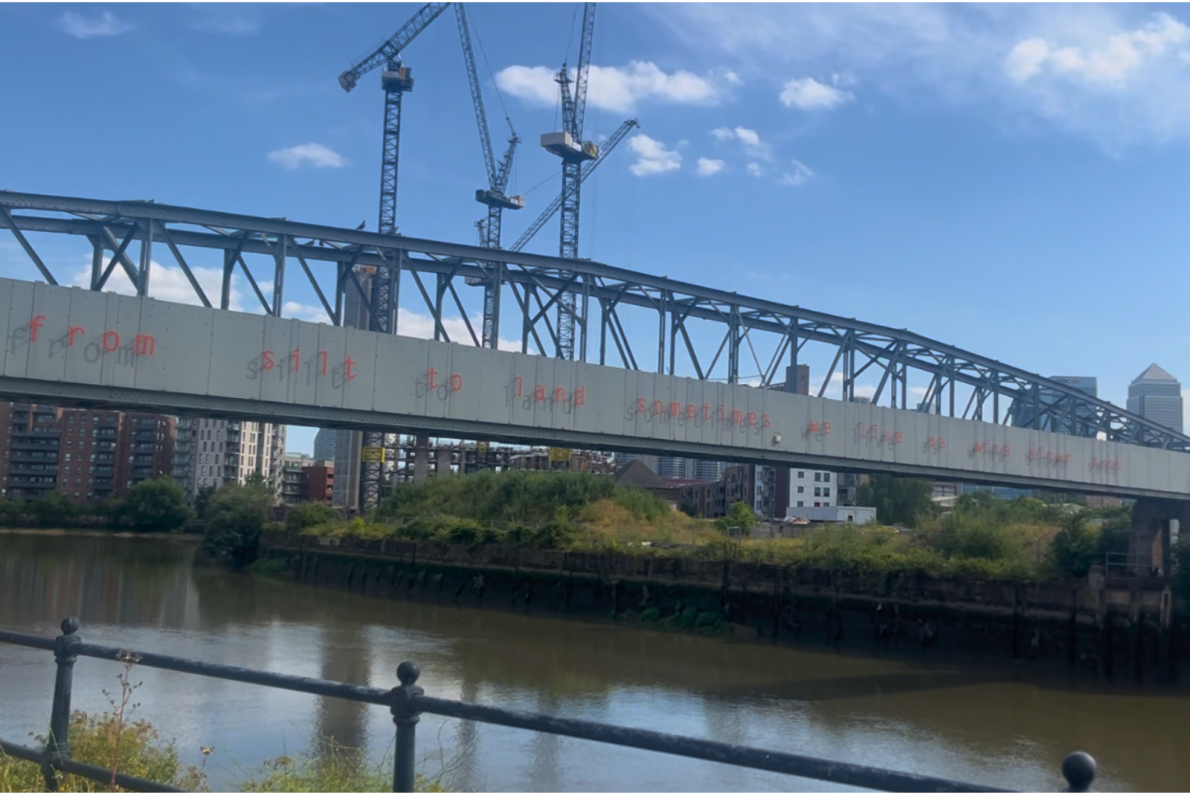 Steel bridge with red text spans a river, silhouetted by cranes and modern buildings. Scene under a clear blue sky conveys an urban, industrial vibe.