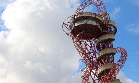 Red steel observation tower with a spiral slide against a partly cloudy sky, conveying a sense of modern architecture and adventure.