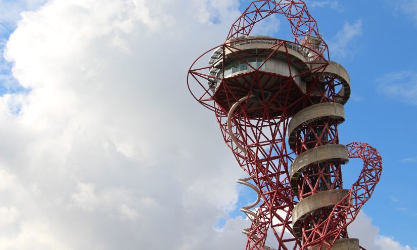 ArcelorMittal Orbit_Photo by Fred Romero Red steel observation tower with a spiral slide against a partly cloudy sky, conveying a sense of modern architecture and adventure.